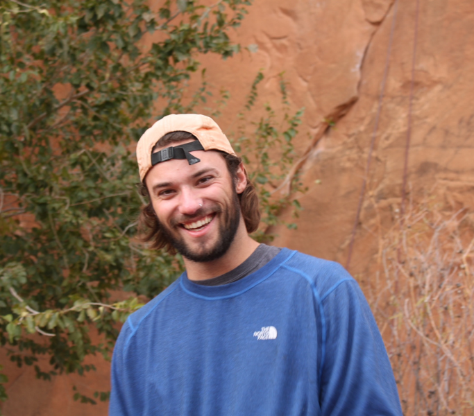 Justin Graham, co-founder of Indigo, smiling and wearing a backwards orange hat and long-sleeve blue t-shirt with orange rocks and a green plant in the background.