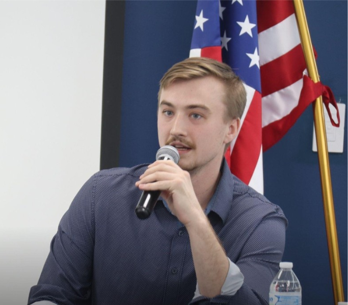 Brandt Bridges speaking into a microphone with an American flag behind him.
