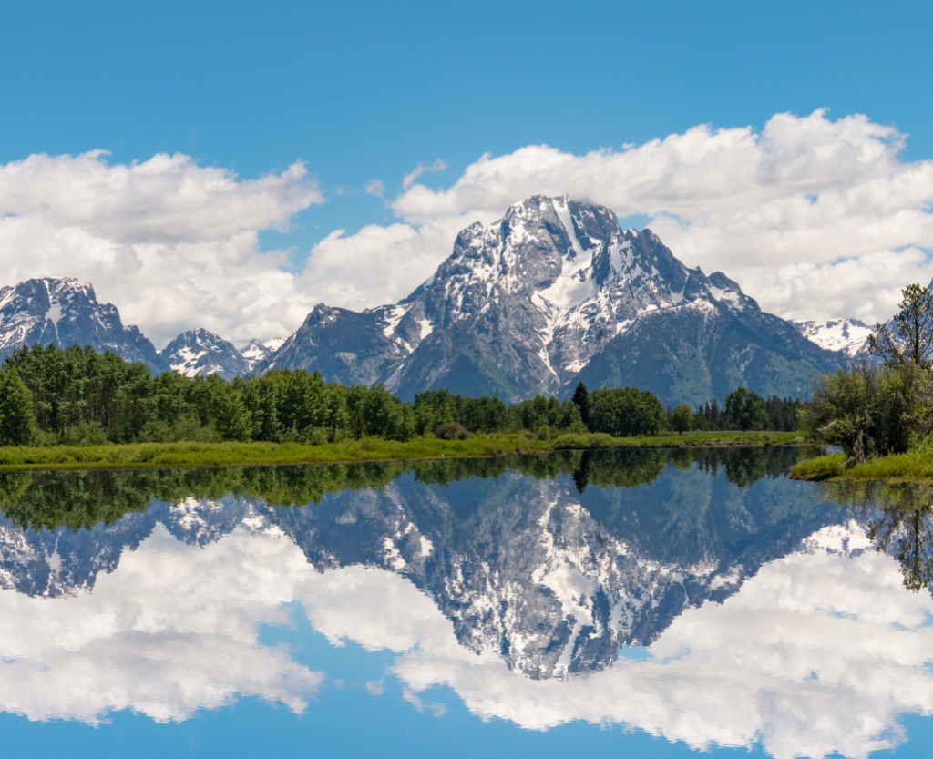 A large mountain with some snow reflected clearly in a lake with a strip of grass and trees between the mountain and the reflection. Blue sky with some clouds behind the mountain.